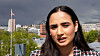 Kvinne foran byutsikt med bygninger, trær og et flagg under overskyet himmel.