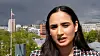 Kvinne foran byutsikt med bygninger, trær og et flagg under overskyet himmel.