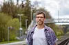 Mann i mønstret skjorte står ved jernbanespor med bro og trær i bakgrunnen.