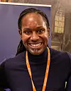 Smilende person med oransje adgangsbånd foran en lilla bakgrunn og et vindusmotiv.