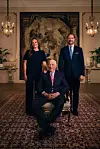 Tre personer i formelle klær poserer i et rikt dekorert rom, med Kong Harald sittende foran Kronprins Haakon og prinsesse Ingrid Alexandra stående.