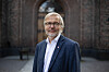 Mann med briller og mørk dress foran en historisk murbygning.
