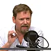 Skjeggete mann i hvit skjorte snakker ved en studiomikrofon.