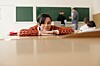 Elev med rødmønstret genser hviler haken på hendene ved en pult i et klasserom.