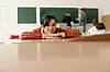 Elev med rødmønstret genser hviler haken på hendene ved en pult i et klasserom.