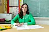 Smilende lærer sitter ved et skrivebord foran en tavle i et klasserom.