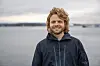 Smilende mann i mørk jakke foran grått vann og overskyet himmel.