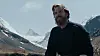 Skjeggete mann i svart genser foran snødekte fjell under blå himmel.